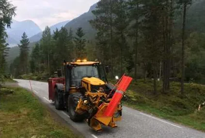 Traktor med redskap foran, på en smal vei mellom skog og fjell